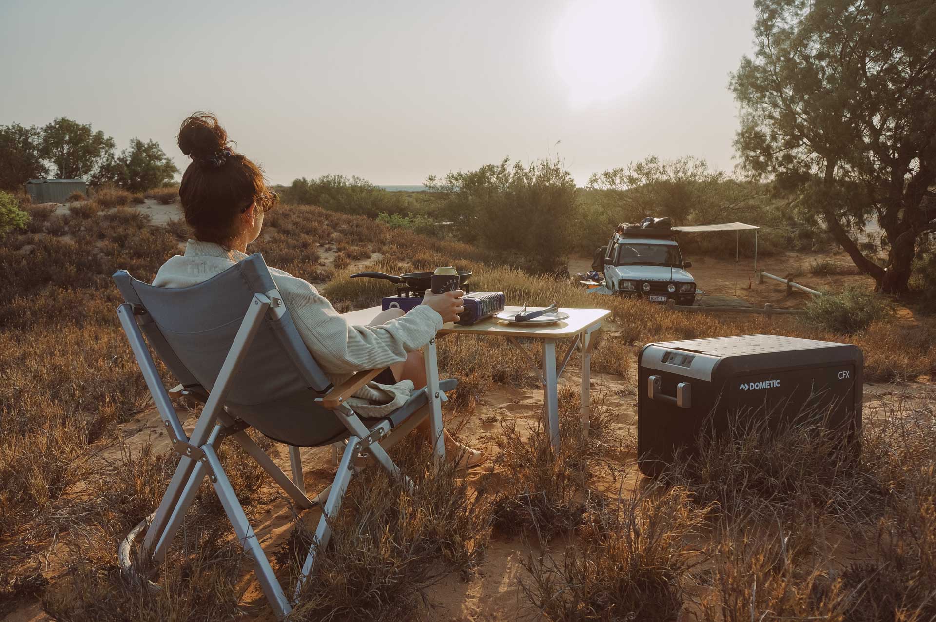 How a Dometic Fridge/Freezer Will Take Your Camping Set Up to the Next Level, gear, dometic, camping, camp cooking, behind shot of a lady on a camp chair looking cooking on a portable stove with a dometic fridge
