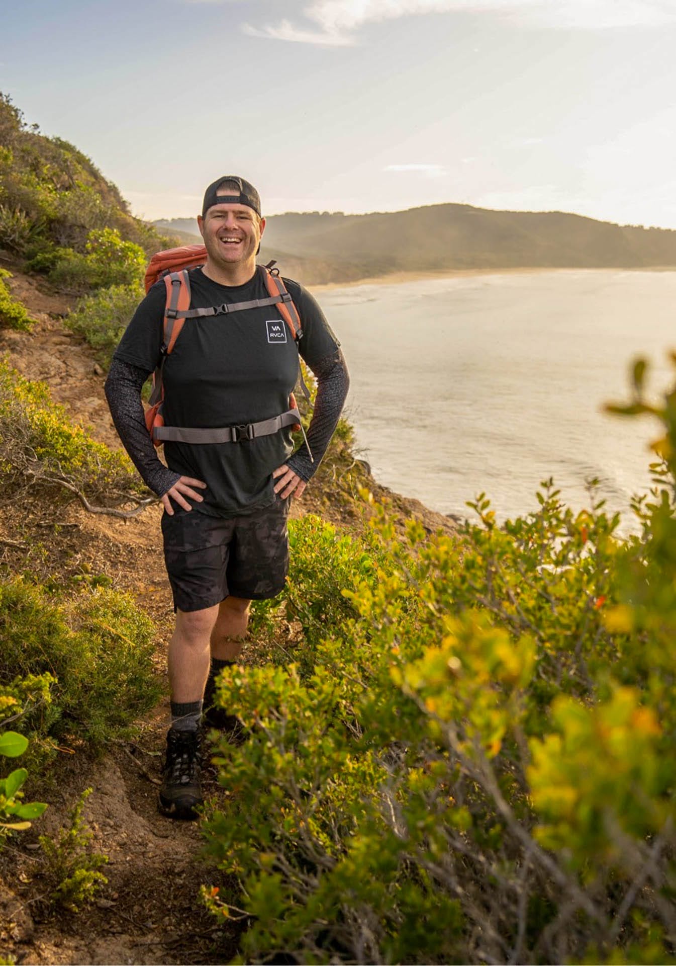 How To Stay an Adventurous Parent and Still Be Home in Time for Family Dinner, microadventures, family adventures, ben dawson wearing an orange backpack while on a coastal headland with bay in the background