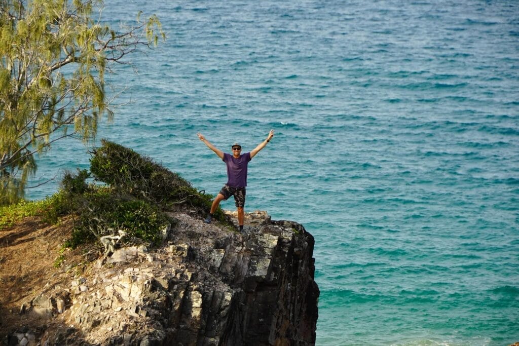 Josh on cliff, by Unknown Photographer, ocean view, coastal adventure, rocky landscape, arms outstretched, peace sign, exploring, travel, risk management