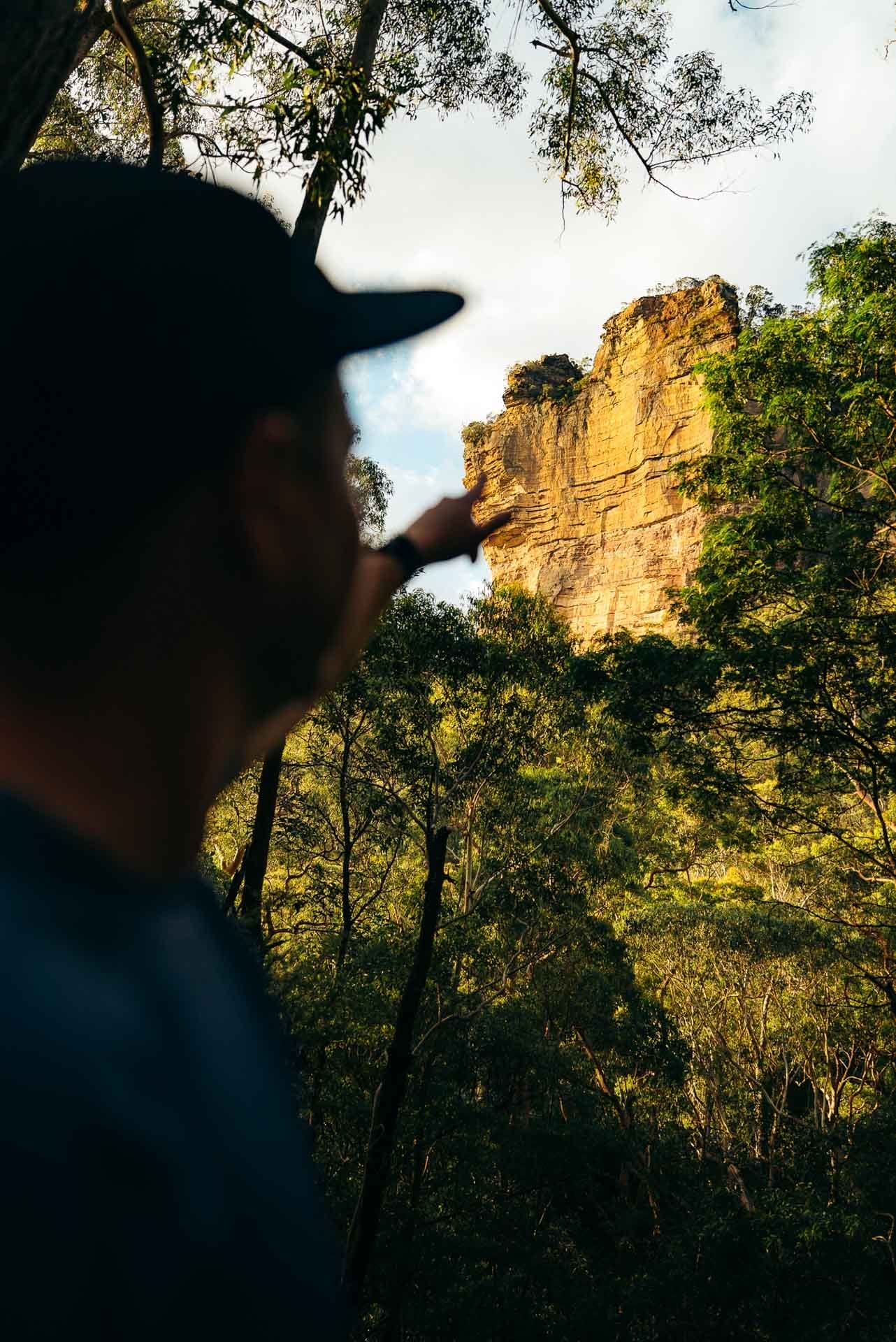 Tim pointing, by Paul, rocky cliff, forest, sunlight, outdoor adventure, escarpment