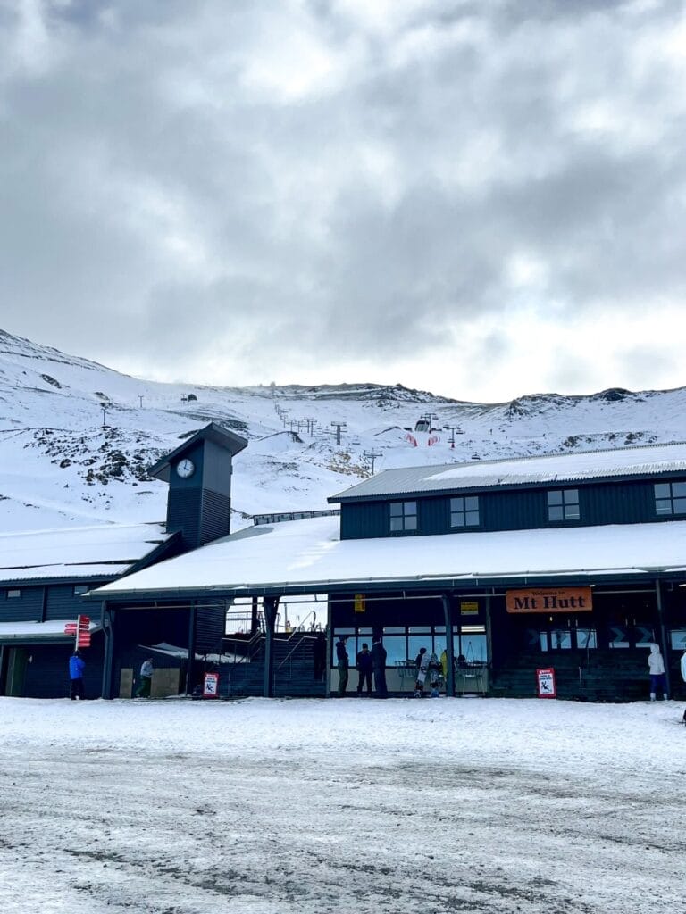 Mt Hutt base lodge, by Tim Whale, snow-covered building, ski lifts, New Zealand ski field, alpine mountains, cloudy sky, clock tower